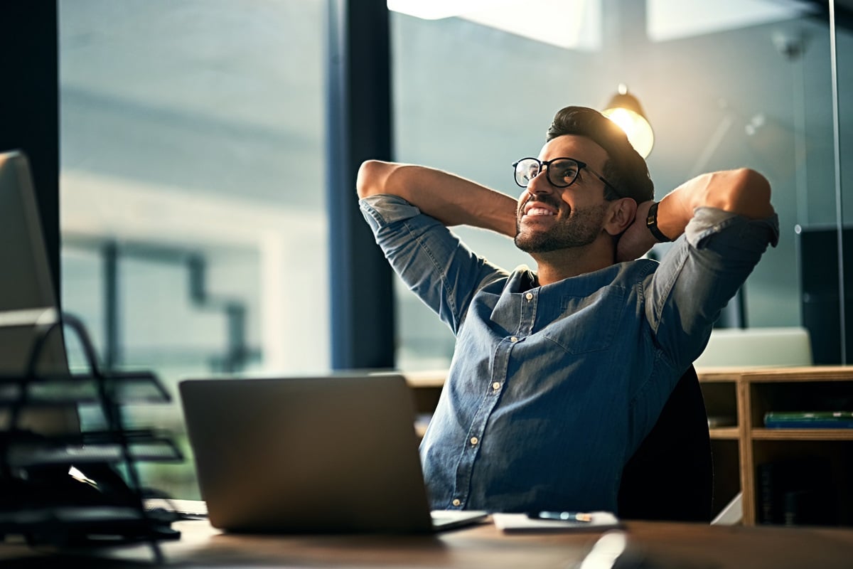 Man sitting at desk with arms behind head looking happy 