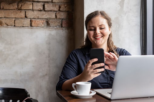 Lady in coffee shop with laptop open, phone in hand smiling 