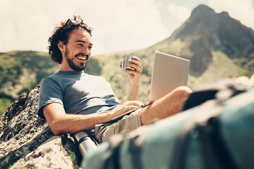 Man laying on rock looking at laptop with coffee