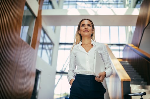 Blonde lady in white shirt walking through office with laptop under her arm