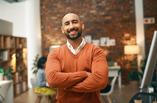 Man in orange smart jumper standing in office smiling with arms folded