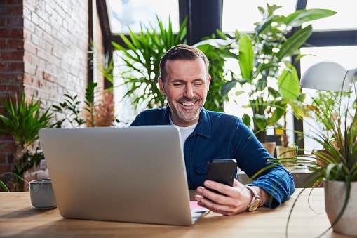 Man sitting in from of laptop, smiling at his phone with plants behind him